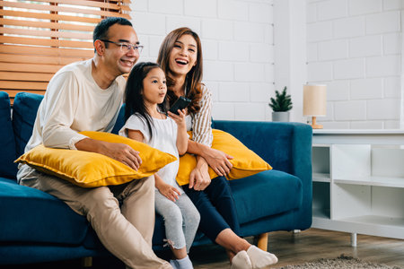 In their modern grooved house a happy Asian family enjoys quality family time watching TV together on the sofa. The father mother son and daughter are all smiles radiating joy.の写真素材