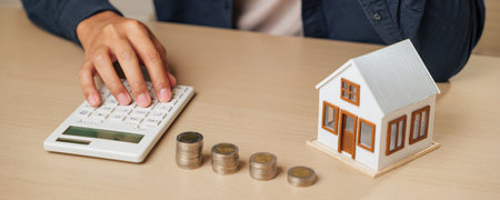 Hands using a calculator with a house model, coins, and cash nearby. Depicts tax analysis, property...