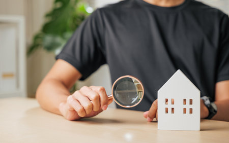 Hand with a magnifying glass examines a cottage house for real estate purposes. Highlights property appraisal, home inspection, and search for a dream house or estate investment opportunities.の写真素材