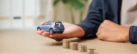 A person holds a blue toy car model near stacked coins of increasing size. Represents car finance concepts like credit, cash payments, loan options, and installment costs for auto purchases.の写真素材
