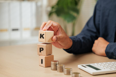 A close-up of a businessman hand holding wooden cube blocks spelling KPI near coins and a calculator. Represents KEY PERFORMANCE INDEX planning, employee performance tracking, and business financial analysis.の写真素材