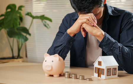 A worried man sits with hands on his forehead near a piggy bank, coins, and a house model. Reflects financial stress, budgeting problems, money risks, and property challenges in banking and finance.の写真素材