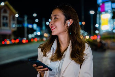 Woman enjoying music through earphones connected to her mobile phone while walking on a night city street. Wireless technology brings people closer. Portrait of a happy girl.の写真素材