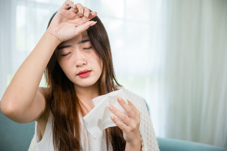 Sick woman sitting under blanket on sofa and sneeze with tissue paper in living room, Asian young female blowing nose sneezing in tissue at home, fever caught coldの写真素材