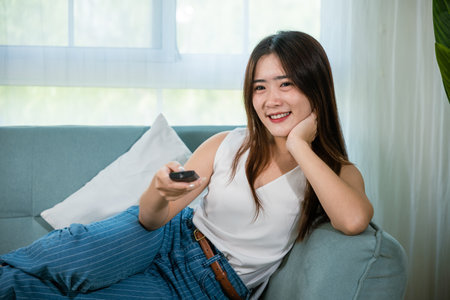 Happy female fun comedy video holding remote watching television, Asian young woman smiling sitting relaxing watch TV holding remote control on sofa in living room, Activity lifestyles conceptの写真素材
