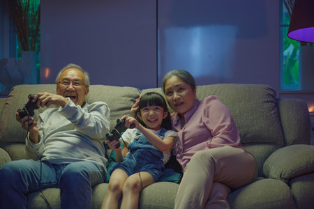 Happy Asian family bonding over a video game on a cozy couch, little girl playing video games console with senior grandparents together with controller joysticks while sitting on the sofa at nightの写真素材