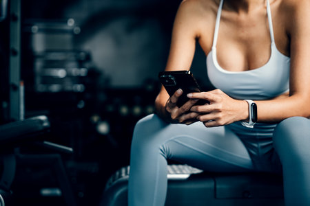Asian woman resting on bench and checking all updates after exercising in gym interior, female using...