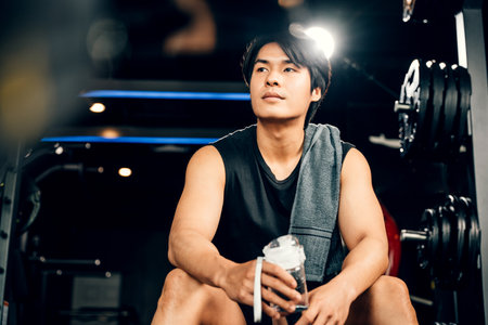 Portrait of Asian young man trained body in black sportswear sitting for rest holding water or protine in fitness gym, Weight training, healthy lifestyle conceptの写真素材