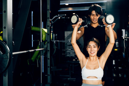 Sporty man and woman exercising with dumbbells, working on their muscle definition and wellbeing under the supervision of their coach, technique of exercise in gym dark gym backgroundの写真素材