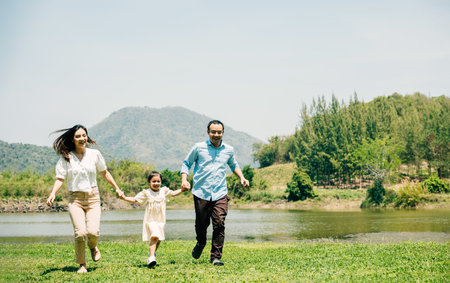 Asian family father mother and daughter holding hands and running lake in park, lifestyle parents and child kid enjoying outdoors together in nature on summer travel, Family relaxation, Front viewの写真素材