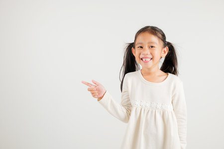 Happy Asian portrait cute young kid girl smiling indicate finger empty space, studio shot isolated on white background, Thai kindergarten child pointing index out with copy spaceの写真素材