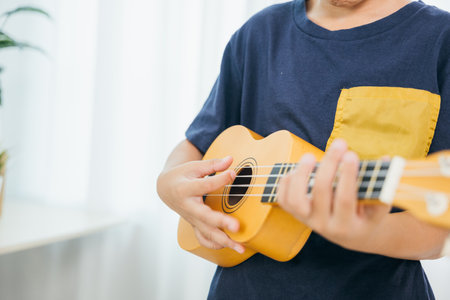 Close-up of a young boys hands in a blue shirt playing a yellow ukulele in the living room. The soft white curtains in the background frame the scene, capturing his passion for music and creativity.の写真素材