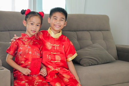 Smiling children in red cheongsam sit on a sofa, holding angpao red envelopes for lucky money. The living room setting highlights family traditions and festive joy during Chinese New Year.の写真素材