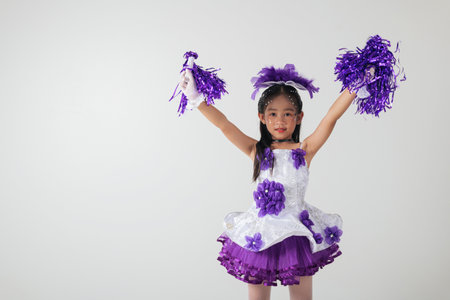 Smiling cheerleader girl wearing purple and white outfit with pom-poms. Studio portrait exuding energy and joy against a white background. cheerleader kidの写真素材