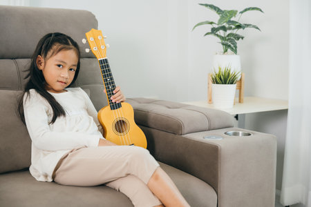 A cheerful girl in a white shirt holds a wooden ukulele while sitting on a grey couch. She looks at the camera, her joy and musical spirit lighting up the modern living room around her.の写真素材