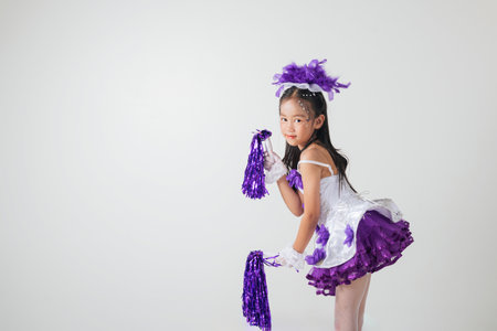 Portrait of a cheerful cheerleader girl in a purple and white outfit, holding purple pom-poms. Smiling brightly in a studio shot against white background. Energetic and festive vibes. cheerleader kidの写真素材
