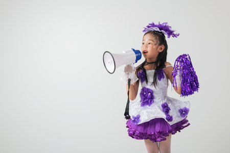 Portrait of a cheerful cheerleader kid in a purple and white outfit, holding a megaphone. Studio shot with a white background, showcasing fun, energy, and team spirit.の写真素材