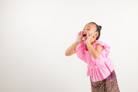 Portrait Thailand kid girl smiling traditional Thai dress costume use hand covering her mouth, studio shot isolated white background, engaged in lively conversation, making announcementの写真素材