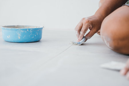 Grouting tiles with a rubber trowel, a hand spreads paste between ceramic tiles on the floor. Tile...