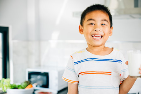 Cheerful Asian little boy holds milk in kitchen, smiling happily. Portrait of cute son enjoying drink. Joyful child sips calcium-rich liquid, radiating happiness at home give me.の写真素材