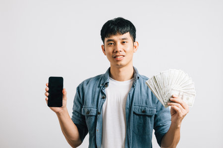Portrait smiling Asian young man holding a bunch of money and mobile phone studio shot isolated white background, happiness and excitement man in dollar banknotes with smartphone blank screenの写真素材