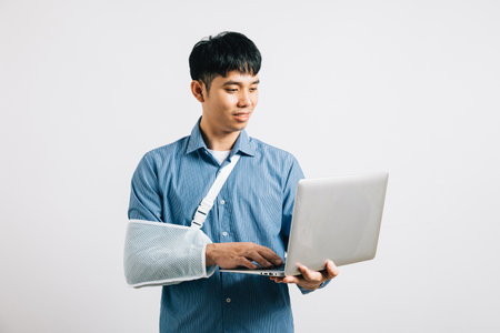 A resilient Asian businessman, with a broken arm in plaster, remains committed to work, holding a laptop. Studio shot isolated on white, emphasizing dedication and recovery.の写真素材