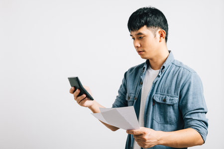 A young man, isolated on a white background, holds a paper and calculator, displaying signs of sadness, tiredness, and strain. He is deeply worried about the bill he is calculating.の写真素材