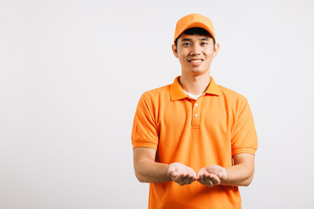 Portrait Asian smiling young delivery man showing something with empty open palm hands studio isolated white background, Happy lifestyle men carry person in an orange shirt uniformの写真素材