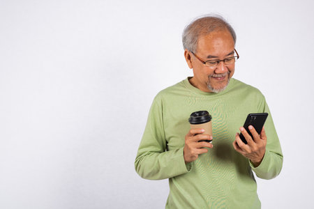 Portrait Asian old man while holding a cell phone and a cup of coffee studio shot isolated on white background. Concept of relaxation and enjoyment, elderly man is enjoy his coffee and check his phoneの写真素材