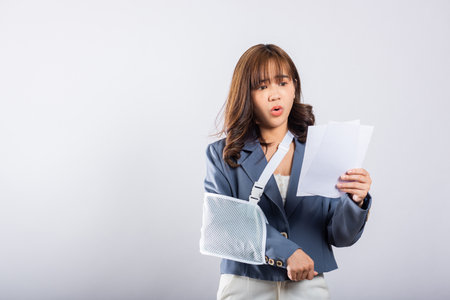 Upset businesswoman with arm in a splint after an accident, finds comfort in sling support while holding medical bills. Asian woman on white background, Broken armの写真素材