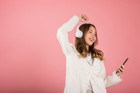 Woman excited smiling listening to music radio in bluetooth headphones and raise hand dancing on smartphone studio shot isolated white background, Happy Asian young female online app in mobile phoneの写真素材