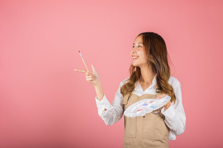Happy female painting using paintbrush and palette with colors, Asian beautiful young woman artist holding brush and paint palette, studio shot isolated on pink background, Paintings and art equipmentの写真素材