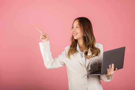 Portrait business woman holding laptop point finger empty space studio shot isolated on pink background, Young smiling happy fun attractive cheerful skilled female hold computer with copy spaceの写真素材