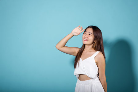 Female raising arm and hand in protection and block sunshine in studio shot isolated on blue background, Asian young woman smile hand up cover face protect sun light and UV, sunscreen cream advertiseの写真素材