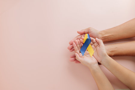 Adult and child hands hold a yellow-blue ribbon on pink background for Down Syndrome awareness. Advocating for Autism Awareness and highlighting Down Syndrome globally on this special day.の写真素材