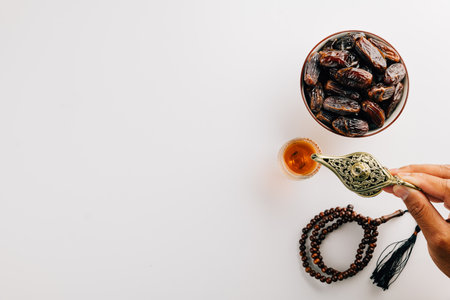Hand pouring tea from a teapot with dates and prayer beads on white. Ramadan celebration highlights holy traditions with a serene flat lay arrangement featuring tea and unity.の写真素材