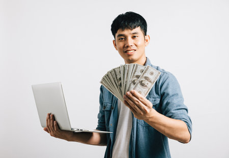 Portrait Asian smiling young man holding laptop computer and fan of money cash studio shot isolated white background, Happy excited lifestyle businessman hold money in dollar banknotesの写真素材