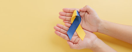 Down Syndrome Day. Yellow and blue awareness ribbon gently held by hands on yellow background. Supporting global efforts for Down Syndrome and Autism Awareness.の写真素材