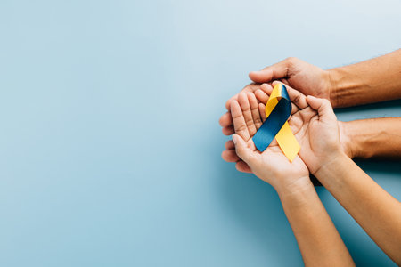 Hands hold a yellow-blue ribbon on a blue background for Down Syndrome advocacy. Promoting Autism Awareness while emphasizing global recognition of Down Syndrome support.の写真素材