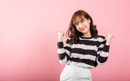 Portrait of a victorious Asian woman with raised fists, celebrating her triumph with a cheerful smile. Studio shot isolated on pink, radiating happiness and success.の写真素材