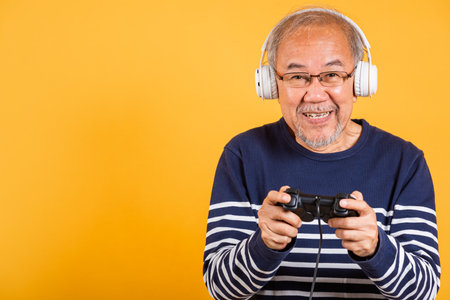 Portrait Asian smiling old man with headphones holding game controller studio shot isolated yellow background, senior man pensioner feeling happy playing a video game with joystick consoleの写真素材