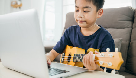 Close-up of young boy hand strumming a ukulele with a laptop displaying an online class nearby. The scene emphasizes modern music learning at home in a relaxed and creative atmosphere.の写真素材