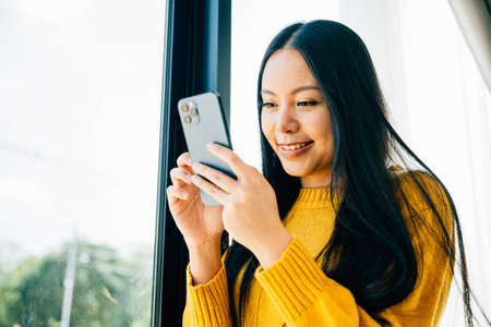 Highlighting smartphone interaction, Woman by the window uses a cellphone for social media texting and connecting at home. Reflecting communication and online reading.の写真素材