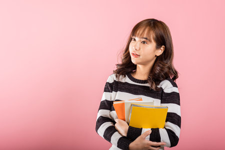 Portrait of beautiful Asian young woman teenage smiling hugging books, female person holding book multicolor, studio shot isolated on pink background with copy space, education conceptの写真素材