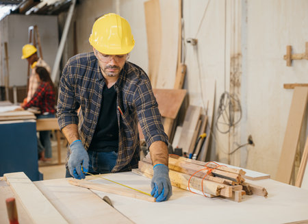 Carpenter wearing a yellow hard hat and safety glasses measuring wood with tape measure. Ideal for woodworking, furniture making, and cabinetry, showcasing precision.の写真素材