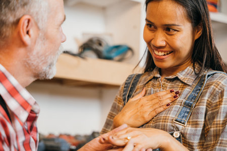 In a wood workshop, a carpenter man presents a ring to a woman carpenter, celebrating their connection. A laptop and small cardboard house sit on the table, symbolizing shared dreams and craftの写真素材
