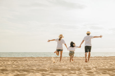 Happy Asian parents with their daughters enjoying playful at beach. Little girls with their mother and father holding hand of child running and pretending on sand. Positive family outdoor activityの写真素材