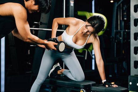 Trainer man teaching woman lifting dumbbell in fitness gym, Happy Asian coach training young female help with a difficult exercise at personal gym, Weight training healthy lifestyle conceptの写真素材