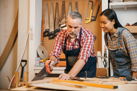 Senior carpenter guides a woman in using a hand saw to cut wood in a workshop. They work together on a woodworking project, surrounded by tools and wooden materials in a well-equipped spaceの写真素材