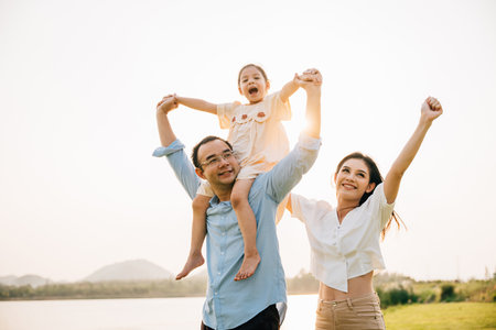 Asian family child girl piggyback riding back on father raising hands like flying outdoor nature park with mother, Happy parent daughter having fun together, Tourist lifestyle family leisure activityの写真素材